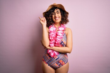 Young beautiful arab woman on vacation wearing swimsuit and hawaiian lei flowers with a big smile on face, pointing with hand and finger to the side looking at the camera.