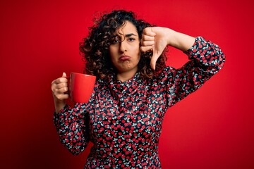 Young beautiful curly arab woman drinking mug of coffee over isolated red background with angry...