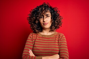 Young beautiful curly arab woman wearing casual striped sweater standing over red background skeptic and nervous, disapproving expression on face with crossed arms. Negative person.