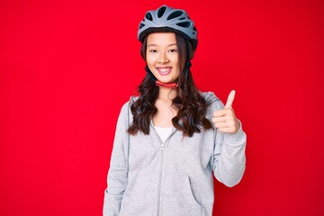 Young beautiful chinese girl wearing bike helmet smiling happy and positive, thumb up doing excellent and approval sign