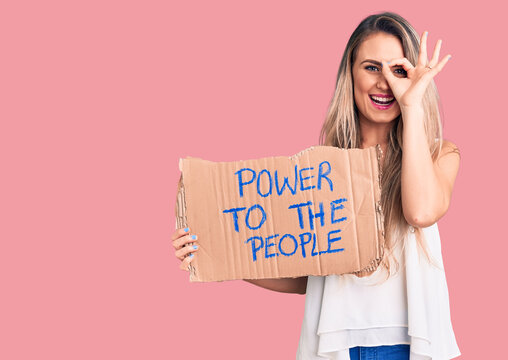 Young beautiful blonde woman holding power to the people cardboard banner smiling happy doing ok sign with hand on eye looking through fingers