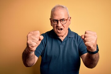 Middle age handsome hoary man wearing casual polo and glasses over yellow background angry and mad raising fists frustrated and furious while shouting with anger. Rage and aggressive concept.