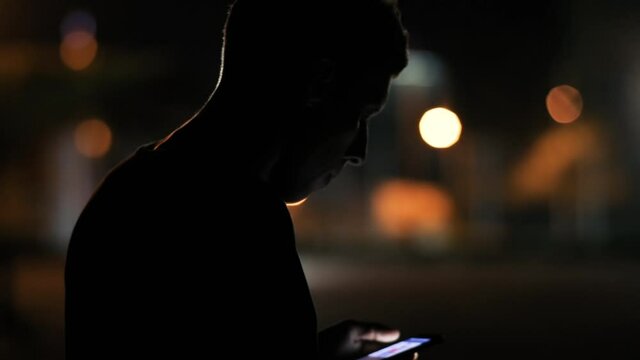 At night, young man texting message on smartphone browsing application news staying in city center street at night. People and nomophobia concept.