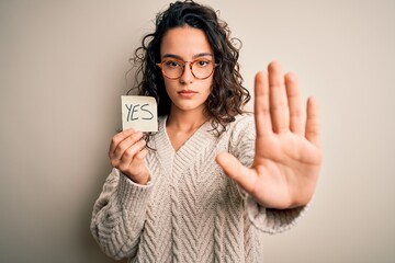 Young beautiful woman with curly hair holding reminder paper with yes word message with open hand...
