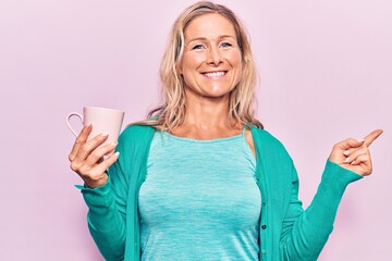 Middle age caucasian blonde woman drinking a cup of coffee smiling happy pointing with hand and finger to the side