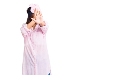 Young beautiful chinese girl wearing sleep mask and pajama covering eyes with hands and doing stop gesture with sad and fear expression. embarrassed and negative concept.