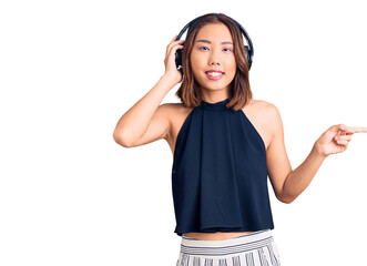 Young beautiful chinese girl listening to music using headphones smiling happy pointing with hand and finger to the side