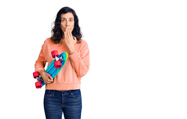 Young beautiful hispanic woman holding skate covering mouth with hand, shocked and afraid for...