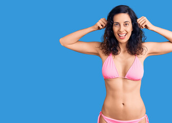 Naklejka premium Young beautiful hispanic woman wearing bikini smiling pulling ears with fingers, funny gesture. audition problem