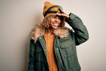 Young african american skier woman with curly hair wearing snow sportswear and ski goggles very happy and smiling looking far away with hand over head. Searching concept.