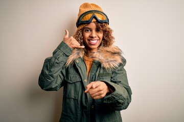 Young african american skier woman with curly hair wearing snow sportswear and ski goggles smiling doing talking on the telephone gesture and pointing to you. Call me.