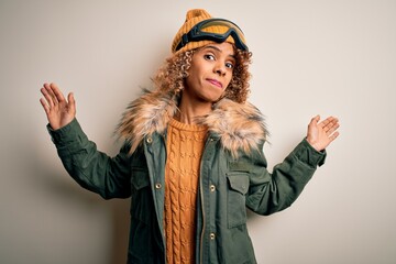 Young african american skier woman with curly hair wearing snow sportswear and ski goggles clueless and confused expression with arms and hands raised. Doubt concept.