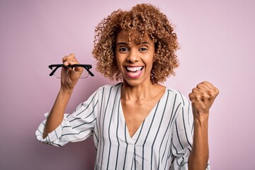 African american optical woman with curly hair holding glasses over isolated pink background screaming proud and celebrating victory and success very excited, cheering emotion