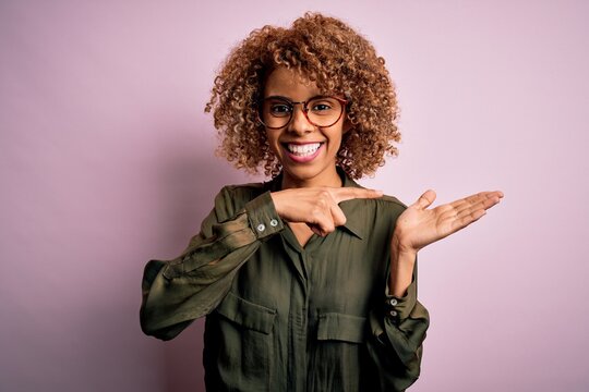 Young beautiful african american woman wearing casual shirt and glasses over pink background amazed and smiling to the camera while presenting with hand and pointing with finger.