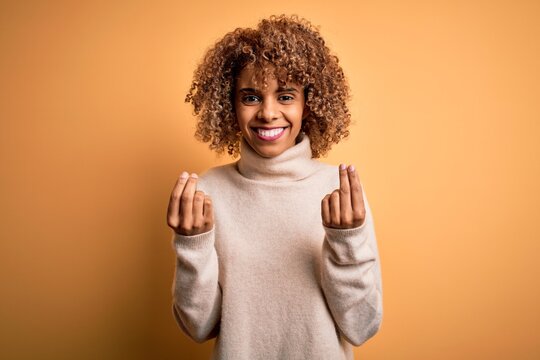 Young beautiful african american woman wearing turtleneck sweater over yellow background doing money gesture with hands, asking for salary payment, millionaire business