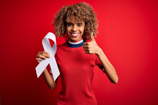 Young african american woman with curly hair holding white cancer ribbon over red background happy with big smile doing ok sign, thumb up with fingers, excellent sign