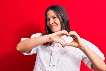 Young beautiful brunette woman wearing casual shirt standing over isolated red background smiling in love showing heart symbol and shape with hands. Romantic concept.