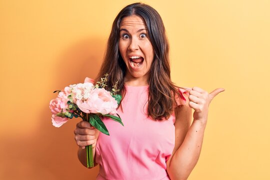 Young beautiful brunette woman holding bouquet of pink flowers over yellow background pointing thumb up to the side smiling happy with open mouth