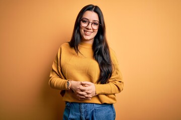 Young brunette woman wearing glasses and casual sweater over yellow isolated background with hands...