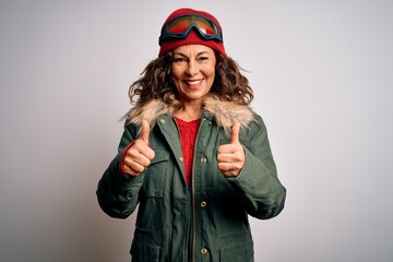 Middle age skier woman wearing snow sportswear and ski goggles over white background success sign doing positive gesture with hand, thumbs up smiling and happy. Cheerful expression and winner gesture.