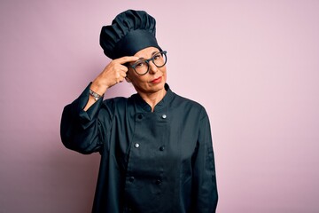 Middle age brunette chef woman wearing cooker uniform and hat over isolated pink background pointing unhappy to pimple on forehead, ugly infection of blackhead. Acne and skin problem