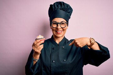 Middle age baker woman wearing cooker uniform and hat holding delicious chocolate cupcake with surprise face pointing finger to himself