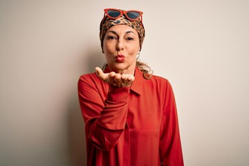 Middle age brunette woman wearing handkerchief on head and shirt over white background looking at the camera blowing a kiss with hand on air being lovely and sexy. Love expression.