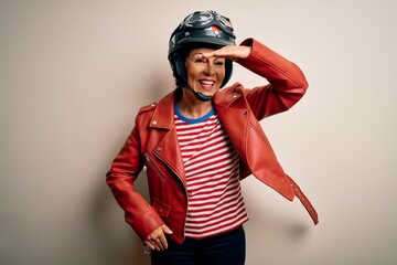 Middle age motorcyclist woman wearing motorcycle helmet and jacket over white background very happy and smiling looking far away with hand over head. Searching concept.