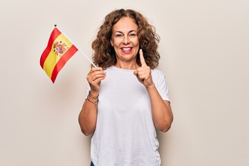 Middle age beautiful patriotic woman holding spanish flag over isolated white background smiling with an idea or question pointing finger with happy face, number one