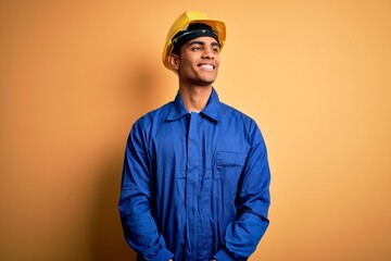 Young handsome african american worker man wearing blue uniform and security helmet looking away to side with smile on face, natural expression. Laughing confident.