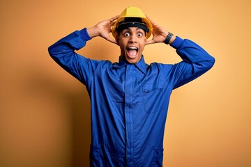 Young handsome african american worker man wearing blue uniform and security helmet Crazy and scared with hands on head, afraid and surprised of shock with open mouth