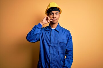Young handsome african american worker man wearing blue uniform and security helmet mouth and lips shut as zip with fingers. Secret and silent, taboo talking