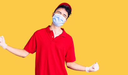 Fototapeta na wymiar Young hispanic man wearing delivery uniform and medical mask very happy and excited doing winner gesture with arms raised, smiling and screaming for success. celebration concept.