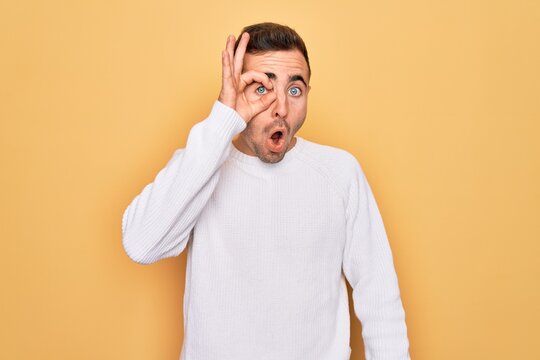 Young handsome man with blue eyes wearing casual sweater standing over yellow background doing ok gesture shocked with surprised face, eye looking through fingers. Unbelieving expression.