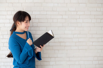 Beautiful asian woman smiling standing thinking and writing notebook on concrete cement white background at home, girl homework on book, education and lifestyle concept.