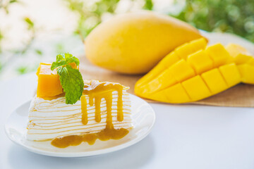 A mango crepe cake sliced on a white plate decorated with a piece of mango fruits and peppermint on a wite table with mango fruit and green nature background.