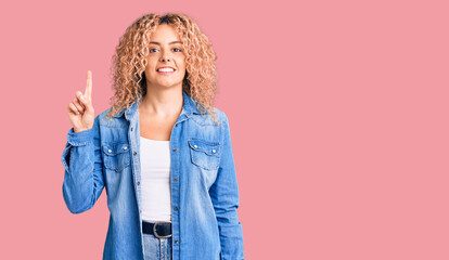 Young blonde woman with curly hair wearing casual denim jacket showing and pointing up with finger number one while smiling confident and happy.