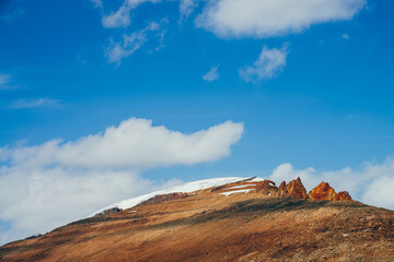 Awesome vivid stony hill with snow in sunlight. Colorful sunny highland landscape with mountain with red crags in top under blue sky. Wonderful alpine scenery with rocks in red orange brown colors.