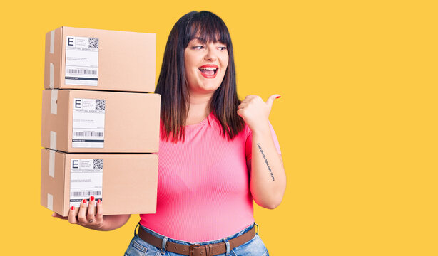 Young plus size woman holding delivery package pointing thumb up to the side smiling happy with open mouth