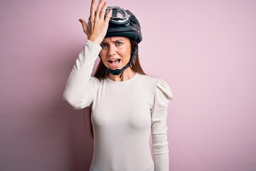 Young beautiful motorcyclist woman with blue eyes wearing moto helmet over pink background surprised with hand on head for mistake, remember error. Forgot, bad memory concept.