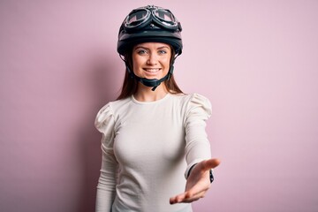 Young beautiful motorcyclist woman with blue eyes wearing moto helmet over pink background smiling cheerful offering palm hand giving assistance and acceptance.