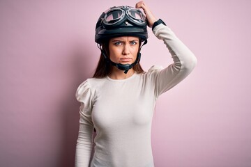 Young beautiful motorcyclist woman with blue eyes wearing moto helmet over pink background confuse and wondering about question. Uncertain with doubt, thinking with hand on head. Pensive concept.