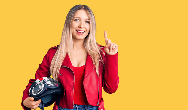 Young beautiful blonde woman holding motorcycle helmet surprised with an idea or question pointing finger with happy face, number one