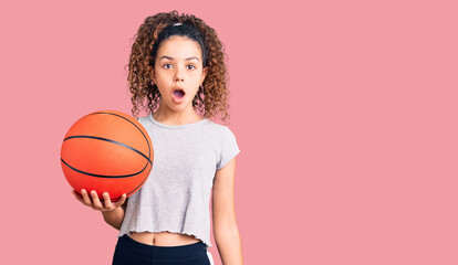 Beautiful kid girl with curly hair holding basketball ball scared and amazed with open mouth for surprise, disbelief face