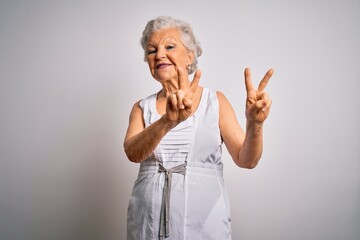 Senior beautiful grey-haired woman wearing casual summer dress over white background smiling looking to the camera showing fingers doing victory sign. Number two.