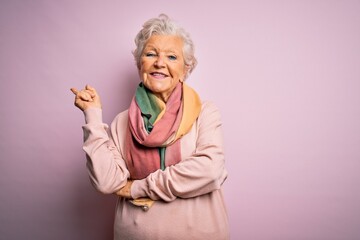 Senior beautiful grey-haired woman wearing casual sweater and scarf over pink background with a big smile on face, pointing with hand and finger to the side looking at the camera.