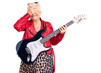 Senior beautiful woman with blue eyes and grey hair wearing a modern look playing electric guitar smiling and laughing with hand on face covering eyes for surprise. blind concept.