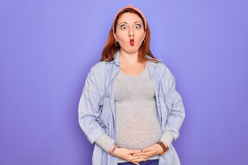 Young beautiful redhead pregnant woman expecting baby over isolated purple background making fish face with lips, crazy and comical gesture. Funny expression.