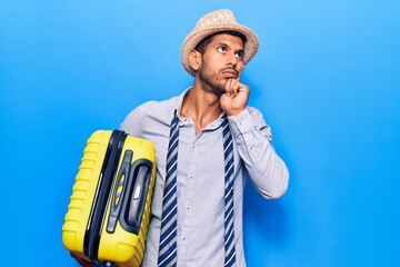 Young latin man wearing summer hat holding cabin bag serious face thinking about question with hand...
