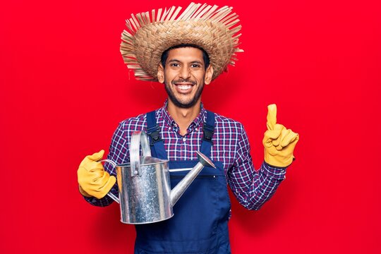 Young latin man wearing farmer hat and gloves holding watering can smiling with an idea or question pointing finger with happy face, number one
